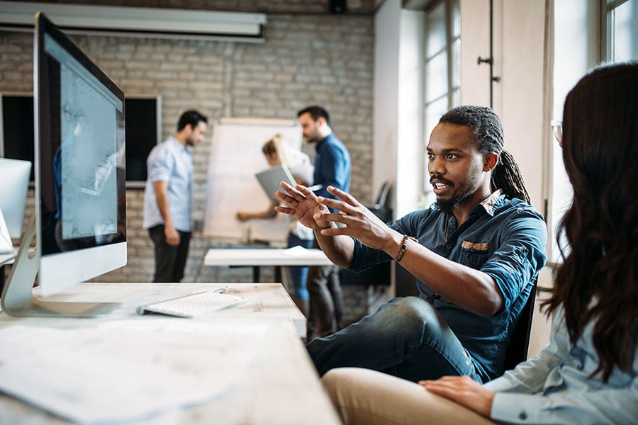 man and woman collaborating with desktop computer at desk with people talking in the background